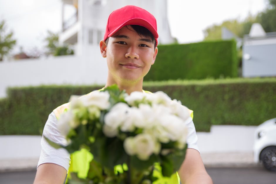 A cheerful male flower delivery courier wearing a red cap and a high-visibility vest stands outdoors, extending a floral bouquet towards the camera. The bouquet, featuring white flowers with lush gree
