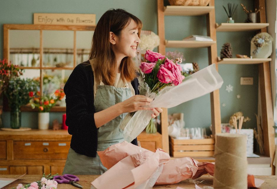 A woman with shoulder-length brown hair, wearing a light green apron over a black top, stands in a flower shop holding a bouquet of fresh pink roses and some smaller filler flowers wrapped in white ti