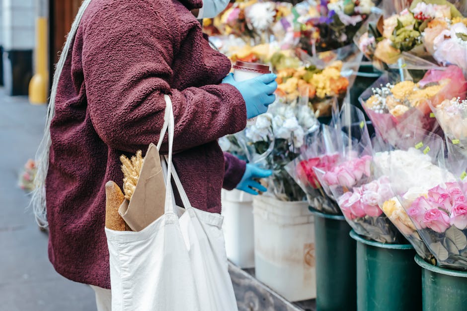 A person wearing a maroon, textured fleece jacket and blue gloves is standing outdoors at a flower stall on a city street, holding a takeaway coffee cup in one hand. They carry a white tote bag with n