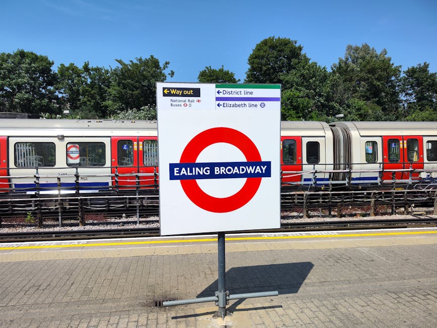 A white sign with a red London Underground circle and blue bar reading 'EALING BROADWAY' is positioned on a train platform in daytime. Behind it, a modern London Overground train with a silver body an