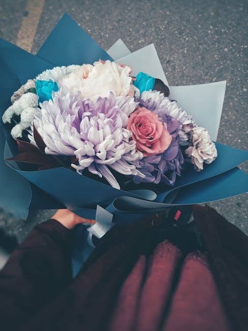 A hand holding a beautifully arranged bouquet of fresh flowers wrapped in layered blue and white paper. The bouquet features a large, soft pink rose at its center, surrounded by white, lavender, and p