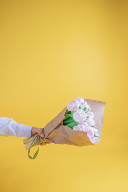 A person wearing a white long-sleeve shirt holds a bouquet of fresh, pastel pink tulips wrapped in light brown craft paper and secured with a green ribbon. The tulips have smooth, rounded petals with 