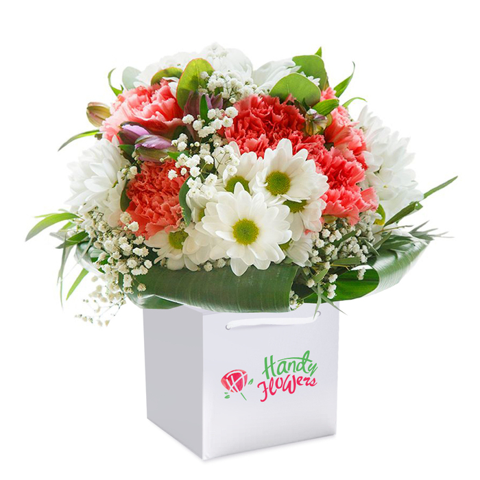 This visually stunning bouquet, designed for floral delivery across Ealing, exudes freshness and thoughtful intent. Set within a spotless white, square gift bag, each bloom is meticulously arranged by experienced hands. Coral and peach carnations, their ruffled petals luxuriously soft, dominate the centerpiece, offering a feast of texture, while brilliant white daisies with striking lime-green centers add an element of pure delight. Gentle touches of white gypsophila are scattered artfully throughout, imparting lightness and a frothy visual texture. The base is ensconced in a collar of broad, glossy green leaves, which envelop the flowers and anchor the composition. Among the lushness, a few purple-tinged buds, possibly alstroemeria, peek out-introducing intrigue and layered depth. Bathed in clean, natural light that highlights each petal's unique beauty, the overall arrangement is round, abundant, and lively, calling to mind the communal feel of Ealing's vibrant gardens and neighbourhood meetups. The white ribbon handles ensure the bouquet is ready for easy pick-up or doorstep delivery along Uxbridge Road. This elegant gift is an ideal way to express heartfelt emotions-offering joy and brightness for birthdays, celebrations, anniversaries, or spontaneous surprises in Ealing.