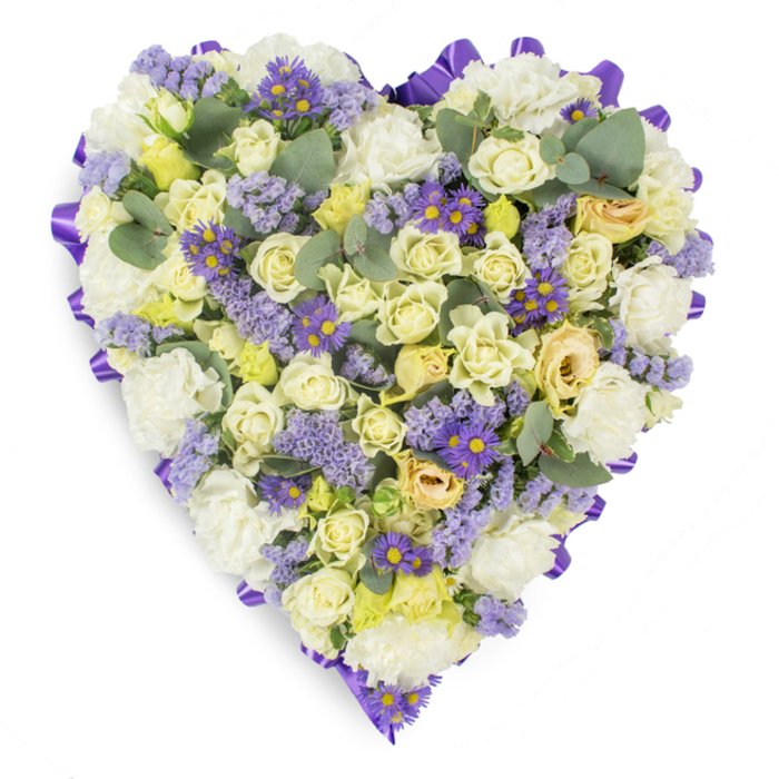 This photograph shows a meticulously arranged, heart-shaped sympathy wreath on a pristine white background, where subtle creams and lavender hues come together in a gesture of remembrance. The arrangement's surface is dominated by clusters of cream and pale yellow roses - some tightly furled, others opening gently and showing soft gradients of lemon and light green - which create a plush, layered texture. Between these roses the classic, ruffled heads of white carnations nestle, adding fullness and a comforting, almost cushion-like quality. Delicate sprigs of lavender-coloured statice and vivid small purple daisy-like asters with golden centres are scattered through the composition, providing bright, cool accents that punctuate the overall serenity and add rhythmic contrast. Blue-green, rounded eucalyptus leaves are placed at intervals to break the floral mass and introduce a silvery-green note and subtle leaf structure. A continuous, rich purple satin ribbon is looped around the entire perimeter, neatly defining the heart's edge and lending the tribute a formal, polished framing. The lighting is soft and even, emphasizing the tactile textures of petals and leaves without creating harsh contrast, suited to studio product photography for memorial floristry. Intended for funerals, wakes or graveside tributes in and around Ealing - from South Ealing to Ealing Common - this composition balances dignity with tenderness, offering a visual and emotional expression of love, remembrance, and peaceful farewell.