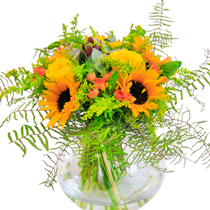 This joyful floral arrangement instantly fills the room with light and warmth, reminiscent of an afternoon wandering around Ealing's stately avenues in high summer. At the heart of the bouquet, dazzling sunflowers with velvet-dark centers and golden petals radiate outwards, catching and reflecting every hint of daylight. Yellow-hued garden roses add a classic, textured richness, their petals curled in intricate spirals that evoke the historic charm of area homes. Glossy red Hypericum berries are scattered throughout the bouquet, their jewel-like brilliance offering playful contrast amid a sea of soft greens and yellows. Flowing ferns, airy solidago, and slender spikes of filler create a harmonious synergy of movement, their shades recalling the rich, ever-changing greens of Pitshanger Park. The subtle blue-green roundness of eucalyptus leaves introduces a gentle, modern element, peeking through with quiet confidence. Resting in a perfectly clear spherical glass vase, the bouquet's green stems glisten in water, allowing the composition's artistry and freshness to shine through. With a fragrance that whispers of garden walks and open spaces, this arrangement-designed for same-day delivery throughout Ealing-is the ideal companion for milestone moments, joyful gatherings, or simply as an elegant dose of happiness in any local home.